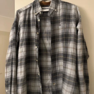 Barbour Skjorta  - Snygg grå rutig skjorta från Barbour i storlek S. Skjortan har klassisk krage med brunt manchesterdetalj, knappar framtill och en bröstficka med Barbour-logga. Materialet är mjuk bomull och passformen är normal. Perfekt för dig som gillar stilrena och tidlösa mönster.