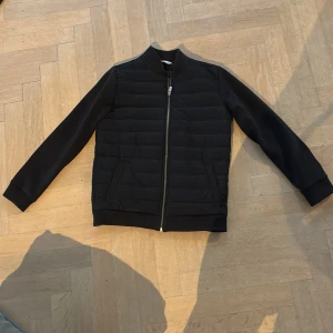 Svart cardigan - Svart bomberjacka med quiltad framsida och rygg, ribbade muddar och krage. Jackan har hel dragkedja framtill och två sidofickor. Materialet är syntet och polyester, vilket gör den lätt och bekväm. Perfekt för dig som gillar stilren och enkel design.