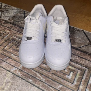 Nike Air Force 1 vita sneakers - Fräscha Nike Air Force 1 sneakers i helvitt med klassisk låg siluett. Skorna har perforerad tå, platt sula och snörning med metall-detalj. Swoosh-loggan på sidan och Nike Air broderat bak. Tillverkade i skinn för en clean och stilren look.