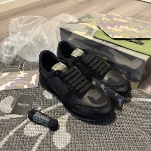 Valentino Garavani Rockrunner sneakers - Svarta Valentino Garavani Rockrunner sneakers med coolt kamouflagemönster. Skorna har snörning, platt sula med nitar och detaljer i mocka och skinn. Märkeslogga på plösen och innersulan. Perfekt för dig som gillar exklusiv streetstyle.
