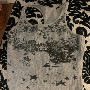 Grå ribbad linnetopp med stjärnor - Säljer en grå ribbad linnetopp med svart grafiskt mönster och stjärnor framtill. Toppen har ärmlös design och rund halsringning, perfekt för dig som gillar edgy och unik stil. Materialet känns mjukt och följsamt mot kroppen.