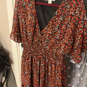 Blommig kortklänning - Säljer en söt kortklänning i storlek 36. Klänningen har ett blommigt mönster i rött, orange, vitt och svart, v-ringad hals och volangdetaljer vid nederkanten. Den har korta, luftiga ärmar och smockad midja som ger en snygg siluett. Perfekt för sommarens alla tillfällen!