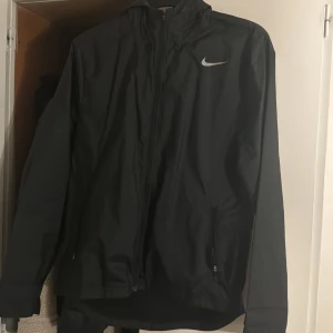 Svart vindjacka från Nike - Svart vindjacka från Nike med stilren design och vit logga på bröstet. Jackan har huva, dragkedja framtill och två sidofickor med dragkedja. Materialet är lätt och syntetiskt, perfekt för blåsiga dagar. Passformen är normal och jackan har långa ärmar.