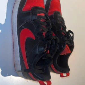 Nike skor svart/röd sneakers - Säljer ett par Nike Jordan 1 sneakers i svart och rött med klassisk röd swoosh och detaljer. Skorna har snörning, rund tå och platt röd sula. Ovandelen är i skinn och textil, med cool kontrast mellan färgerna. Perfekt för dig som gillar streetstyle och ikoniska sneakers.