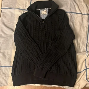 Svart stickad tröja Lindbergh M - Svart stickad tröja från Lindbergh White i relaxed fit. Tröjan har kabelstickat mönster, hög krage och half zip-dragkedja framtill. Perfekt för dig som gillar en chill och stilren look. Materialet är mjukt och håller dig varm under kyliga dagar.