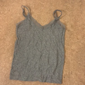 Grå ribbad linnetopp med spets - Grå linnetopp med smala axelband och v-ringning dekorerad med spets. Toppen har ett ribbat mönster och är tillverkad i ett mjukt material som känns bekvämt mot huden. Perfekt att styla med jeans eller kjol för en enkel och snygg look.