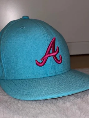 Turkos New Era 59FIFTY keps - Snygg turkos keps från New Era, modell 59FIFTY, med Atlanta Braves A-logga i rosa brodyr framtill och MLB-logga bak. Klassisk formad skärm och stängd baksida. Perfekt för dig som vill sticka ut med färg och sportig vibe. Storlek 7 55,8 cm.