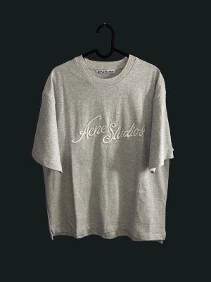 Grå Acne Studios t-shirt - Snygg grå t-shirt från Acne Studios med broderad logga framtill i vitt. Klassisk rund halsringning och avslappnad passform. Perfekt till jeans eller shorts för en clean och trendig look.