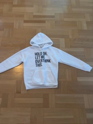 Vit hoodie med tryck - Säljer en vit hoodie med svart texttryck på bröstet där det står 'HOLD ON, LET ME OVERTHINK THIS.' Hoodien har huva med dragsko och en stor magficka. Perfekt för dig som gillar bekväma och stilrena plagg med en rolig twist.
