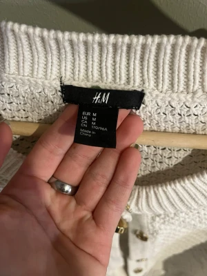 Vit stickad kofta från H&M - Snygg vit stickad kofta från H&M med rund halsringning och dekorativa guldknappar framtill. Koftan har ett grovt stickat mönster och ribbade muddar vid ärmslut och nederkant. Perfekt att slänga över en topp för en chill look.