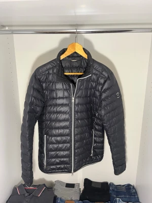 Svart dunjacka från J.Lindeberg - Snygg svart dunjacka från J.Lindeberg med quiltad design och vita dragkedjor. Jackan har en innerficka och schysst nu till vår. Jag är 180 cm lång och väger 76 kg. Hör av dig för fler frågor