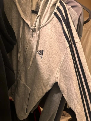 Grå zip hoodie från Adidas / pris går att diskutera  - Snygg grå hoodie från Adidas med dragkedja framtill och klassiska svarta ränder längs ärmarna. Har huva med dragsko och Adidas-logga broderad på bröstet. Perfekt för en sportig och avslappnad stil.