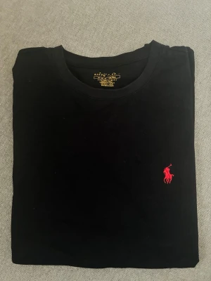 Svart t-shirt från Polo Ralph Lauren - Snygg svart t-shirt från Polo Ralph Lauren med röd broderad logga på bröstet. Modellen är custom slim fit och har klassisk rund hals. Tillverkad i mjuk bomull som känns skön mot huden. Perfekt basplagg för en clean och stilren look.