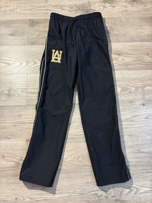 Under Armour - Svarta mjukisbyxor med vita sidoränder och en stor broderad patch med bokstäver på vänster lår. Byxorna har dragsko i midjan och raka ben. Perfekta för en chill och sportig look. Storlek XS passar S❗️