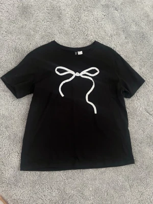 Svart t-shirt med vit rosett - Säljer en svart t-shirt från H&M med en stor vit rosett tryckt på bröstet. T-shirten har korta ärmar och en klassisk rund halsringning. Perfekt för dig som gillar enkla men snygga detaljer.