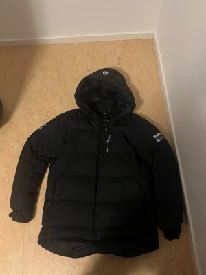 Sail racing jacka - Svart dunjacka från sail racing med huva och dragkedja framtill. Jackan har en quiltad design, logga på ärmen och bröstet samt praktiska fickor. Perfekt för kalla dagar och har en klassisk, stilren look.