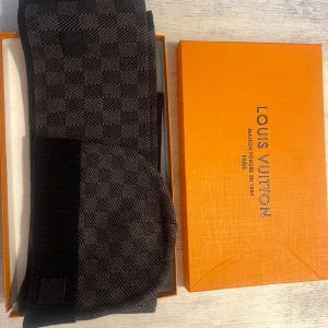 Louis Vuitton mörkgrå halsduk - Snygg mörkgrå halsduk från Louis Vuitton med klassiskt rutmönster och diskret logotyp i läder. Halsduken har en stilren design och är tillverkad i ett mjukt, stickat material som känns lyxigt och bekvämt. Perfekt accessoar för att lyfta din outfit.