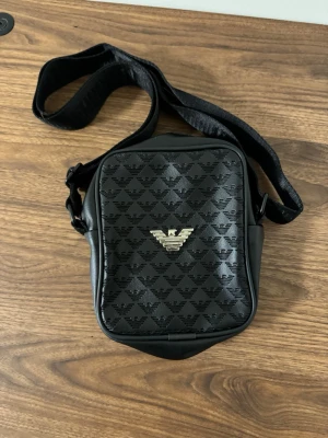 Svart axelväska från Emporio Armani - Snygg svart axelväska från Emporio Armani med justerbar rem. Väskan har präglat logomönster över hela ytan och en silverfärgad logga framtill. Tillverkad i skinnliknande material och har dragkedja baktill för extra förvaring.