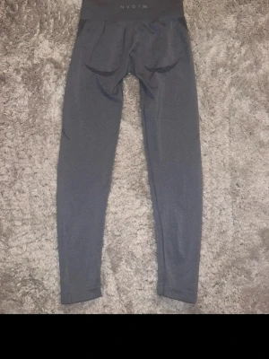 Grå seamless tights från NVGTN - Säljer ett par grå seamless tights från NVGTN med hög midja och diskreta konturdetaljer vid rumpan. Materialet är stretchigt och passar perfekt för gym eller träning. Tightsen har en tight passform och är utan synliga sömmar för extra komfort. På de två sista bilderna finns det en liten ”skada” men inget som syns eller påverkar 