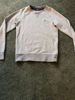 Ljusgrå sweatshirt från Gant - Säljer en stilren ljusgrå sweatshirt från Gant med diskret logga broderad på bröstet. Tröjan har rund halsringning, långa ärmar och breda muddar vid ärmslut och nederkant. Perfekt för en avslappnad och snygg look.