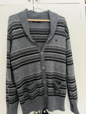 Grå randig kofta från Junk de Luxe - Snygg grå kofta från Junk de Luxe med svarta och vita ränder. Koftan har knäppning framtill, två fickor och en liten broderad logga på bröstet. Tillverkad i mjukt stickat material och har ribbade muddar vid ärmslut och nederkant.