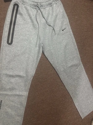 Grå Nike NOCTA mjukisbyxor - Snygga ljusgrå mjukisbyxor från Nike NOCTA med dragsko i midjan och en unik dragkedjedetalj på vänster ben. Byxorna har en tapered passform och små tryckta detaljer på benen. Perfekta för chill eller träning. Helt ny