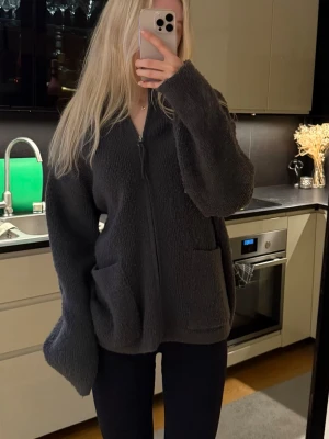 Mörkgrå fleecekofta med dragkedja - Mysig mörkgrå fleecekofta med dragkedja framtill och två stora fickor. Koftan har en avslappnad passform och långa ärmar, perfekt för lager på lager. Materialet är fluffigt och mjukt, vilket gör den extra bekväm. Storlek S-M