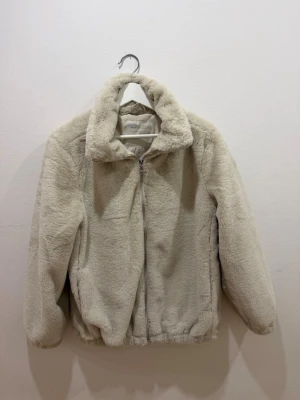 Beige teddyjacka med dragkedja - Mysig beige teddyjacka med hög krage och dragkedja framtill. Jackan har långärmad design och elastisk nederkant för en skön passform. Perfekt för kalla dagar när du vill vara både varm och stylish.