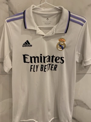 Real Madrid matchtröja Adidas vit 2022/2023 - Snygg vit Real Madrid matchtröja från Adidas med lila detaljer på axlarna och klubbmärke på bröstet. Tröjan har svart Adidas-logga och Emirates Fly Better-tryck framtill. Tillverkad i lätt och funktionellt material, perfekt för fotboll eller träning.