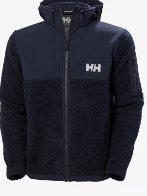 Mörkblå teddyjacka från Helly Hansen - Mysig mörkblå teddyjacka från Helly Hansen med huva och dragkedja framtill. Jackan har HH-logga på bröstet, mjukt teddyfoder och praktisk ficka på ärmen. Perfekt för kyliga dagar och ger en sportig look.