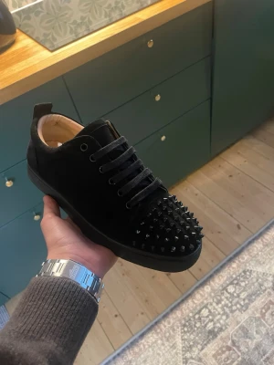 Svarta sneakers med nitar från Louboutin - Svarta sneakers i mocka från Christian Louboutin med coola svarta nitar på tån och klassisk röd sula. Skorna har snörning och en rund tå, vilket ger en edgy och lyxig vibe. Perfekta för dig som vill sticka ut med din stil. A-kopior i bra skick!