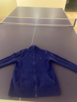 Blå stickad ziptröja från Polo Ralph Lauren - Mörkblå stickad ziptröja från Polo Ralph Lauren med röd broderad logga på bröstet. Tröjan har hög krage, dragkedja framtill och två fickor. Ribbstickade muddar vid ärmslut och nederkant. Perfekt för en chill och stilren look.