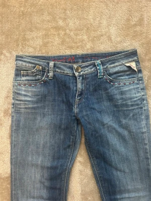Blå bootcut jeans med detaljer - Säljer ett par jeans då de knappt används. Snygga blå bootcut jeans med broderade detaljer i rött och blått vid fickorna. Jeansen har klassisk femficksdesign. Passar mig som är 165 och midjemått är runt 80-81