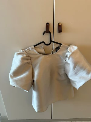 Beige topp med puffärmar Vero Moda - Trendig beige topp från Vero Moda med stora puffärmar och rund halsringning. Toppen har en croppad passform och är tillverkad i ett glansigt material som ger en lyxig känsla. Perfekt för dig som vill sticka ut med en cool och modern look.