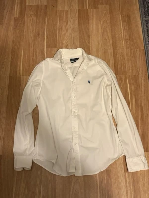 Vit skjorta från Ralph Lauren - Klassisk vit skjorta från Ralph Lauren med blå broderad logga på bröstet. Skjortan har lång ärm, knappar framtill och en tidlös krage. Perfekt till jeans eller chinos för en clean och stilren look. Skjortan är 152 cm och har inga defekter! Skriv ifall att ni har frågor!