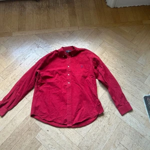 Röd skjorta från Ralph Lauren - Klassisk röd skjorta från Ralph Lauren i bomull med button down-krage och blå broderad logga på bröstfickan. Skjortan har vita knappar och lång ärm, perfekt för en stilren och avslappnad look. Säljer då den är lite stor på mig. XL men passar L jättebra.