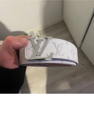  monogram bälte - Snyggt vitt bälte med klassiskt monogrammönster och stor -logga i silver som spänne. Bältet är gjort i präglat läder och har en stilren, modern look som passar till jeans eller byxor.