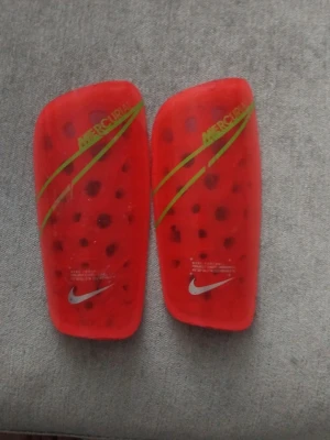 Röda benskydd Nike Mercurial - Säljer ett par röda benskydd från Nike Mercurial-serien. De har svarta prickar och gröna detaljer med Nike-logga i silver. Perfekta för fotbollsträning eller match. Lätta och smidiga att använda det är bra