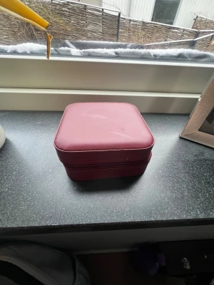 Vinröd smyckesbox i läderimitation - Snygg vinröd smyckesbox med fyrkantig form och dragkedja runt om. Perfekt för att förvara ringar, halsband och andra små accessoarer. Tillverkad i läderimitation med stilren design som passar på sminkbordet eller i väskan.