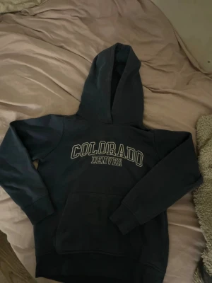 Mörkblå Colorado Denver hoodie - Mörkblå hoodie med stor huva och magficka. På bröstet står det 'COLORADO DENVER' i vita bokstäver. Tillverkad i mjukt och bekvämt material, perfekt för chill dagar. Klassisk college-stil som passar till det mesta.