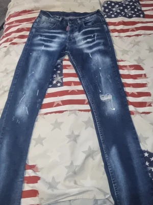 Blå Dsquared2 jeans med slitningar - Säljer ett par blå jeans från Dsquared2 med slitna detaljer, färgstänk och en patch bak i midjan. Jeansen har en smal passform och klassisk femficksdesign. Perfekta för dig som gillar en edgy och trendig look.