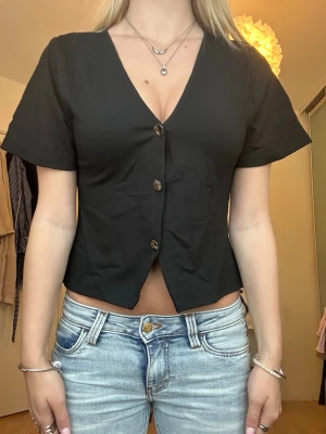 Svart v-ringad blus med knappar - Snygg blus från lager 157 med v-ringning och korta ärmar. Modellen är croppad och har dekorativa knappar framtill. Perfekt till jeans eller kjol för en trendig look.