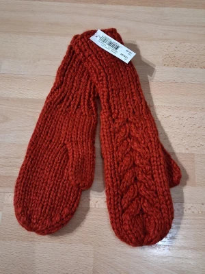 Röda stickade tumvantar - Säljer ett par snygga röda stickade tumvantar med flätmönster på ovansidan. Vantarna är gjorda i tjockt garn och har en klassisk form som passar perfekt för kalla dagar. Märket är Fashion och materialet ser ut att vara ullblandning.