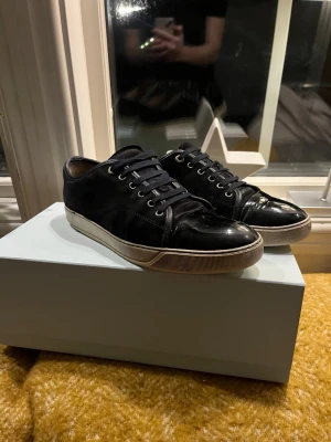 Svarta sneakers från Lanvin med lackdetalj - Snygga svarta sneakers från Lanvin med klassisk snörning och rund tå. Ovandelen är i mocka med glansig lackad tå och vit sula. Perfekta för dig som gillar stilrena och lyxiga detaljer. Märkeslogga på sulan. Behöver tvättas 