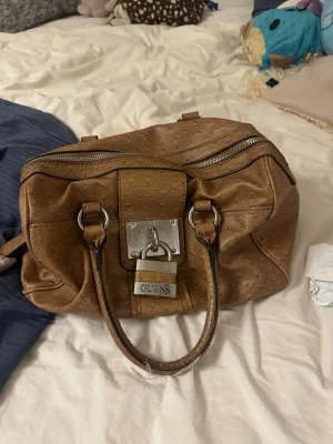 Brun handväska från Guess med lås - Säljer en brun handväska från Guess i syntetmaterial med struktur som påminner om struts. Väskan har dubbla handtag, silverfärgade metalldetaljer och ett stort lås framtill. Insidan är fodrad med mönstrat tyg i vitt och orange med Guess-logga. Fråga innan köp 