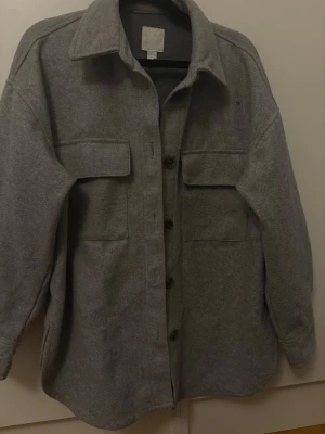 Grå overshirt med fickor och knappar - Stilren grå overshirt med klassisk krage, stora bröstfickor och knäppning framtill. Overshirten har lång ärm och är gjord i ett mjukt material som känns lite ulligt. Perfekt att slänga över en hoodie eller t-shirt för en chill look.