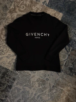 Svart sweatshirt från Givenchy - Svart sweatshirt från Givenchy med vit logotyptext 'GIVENCHY PARIS' på bröstet. Tröjan har rund halsringning, långa ärmar och ribbade muddar. Perfekt för en stilren och trendig look. Det står L på lappen men passar mindre, DM för måttet