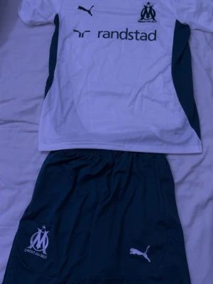Marseille fotbollskit från Puma - Säljer ett Marseille fotbollskit från Puma med vit tröja och mörkblå shorts. Tröjan har klubbmärke, Puma-logga och Randstad som sponsortryck. Shortsen har klubbmärke och Puma-logga. Perfekt för träning eller match!