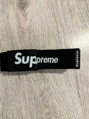 Supreme Bälte  - Snyggt svart bälte från Supreme med tydlig vit logga på både spännet och änden. Bältet är gjort i slitstarkt tygmaterial och har en klassisk, rak form. Perfekt accessoar för att lyfta din streetwear-look.