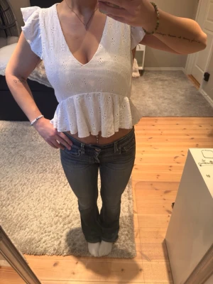 Vit blus med volang och v-ringning - Söt vit blus med v-ringning, broderade detaljer och volang nedtill. Ärmlös modell med små volangärmar och croppad passform. Perfekt till jeans eller kjol för en trendig look.